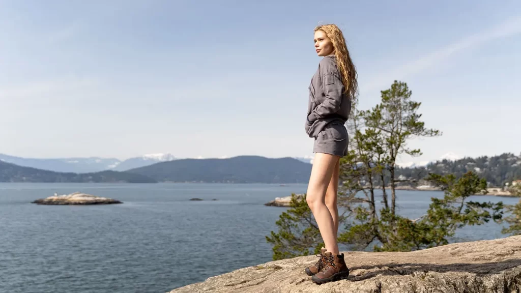 A woman hiking standing looking over an ocean viewpoint wearing and organic cotton hoodie and pocket shorts
