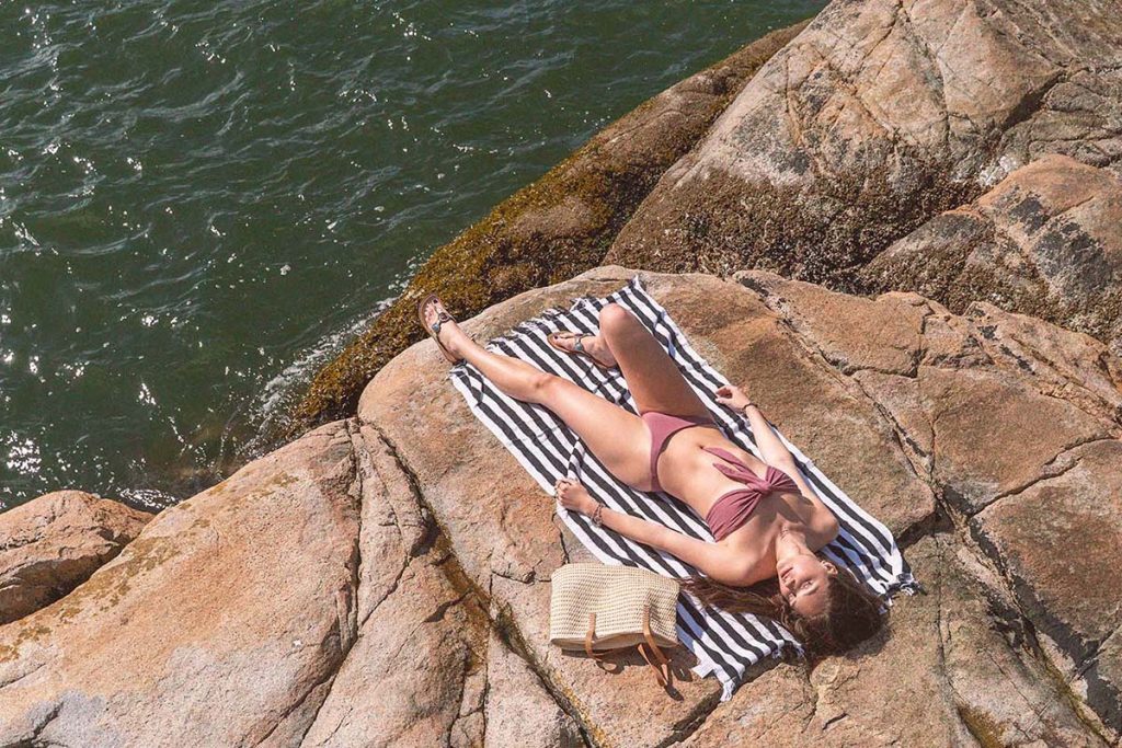 A woman laying on a striped towel on an oceanside rocky shoreline wearing a dusty rose organic bikini made from natural fibers