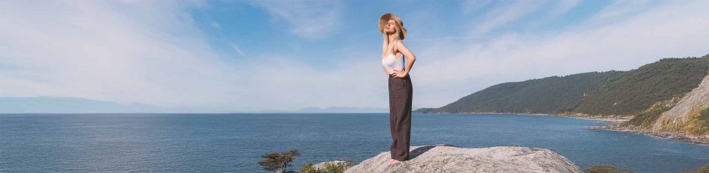 A woman standing on a rock looking over the ocean wearing dark brown linen pants with a white bamboo bodysuit